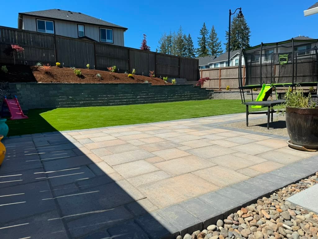 Two-tone paver patio, artificial turf and a tiered block retaining wall with fresh plantings in a family backyard by GI Landscape
