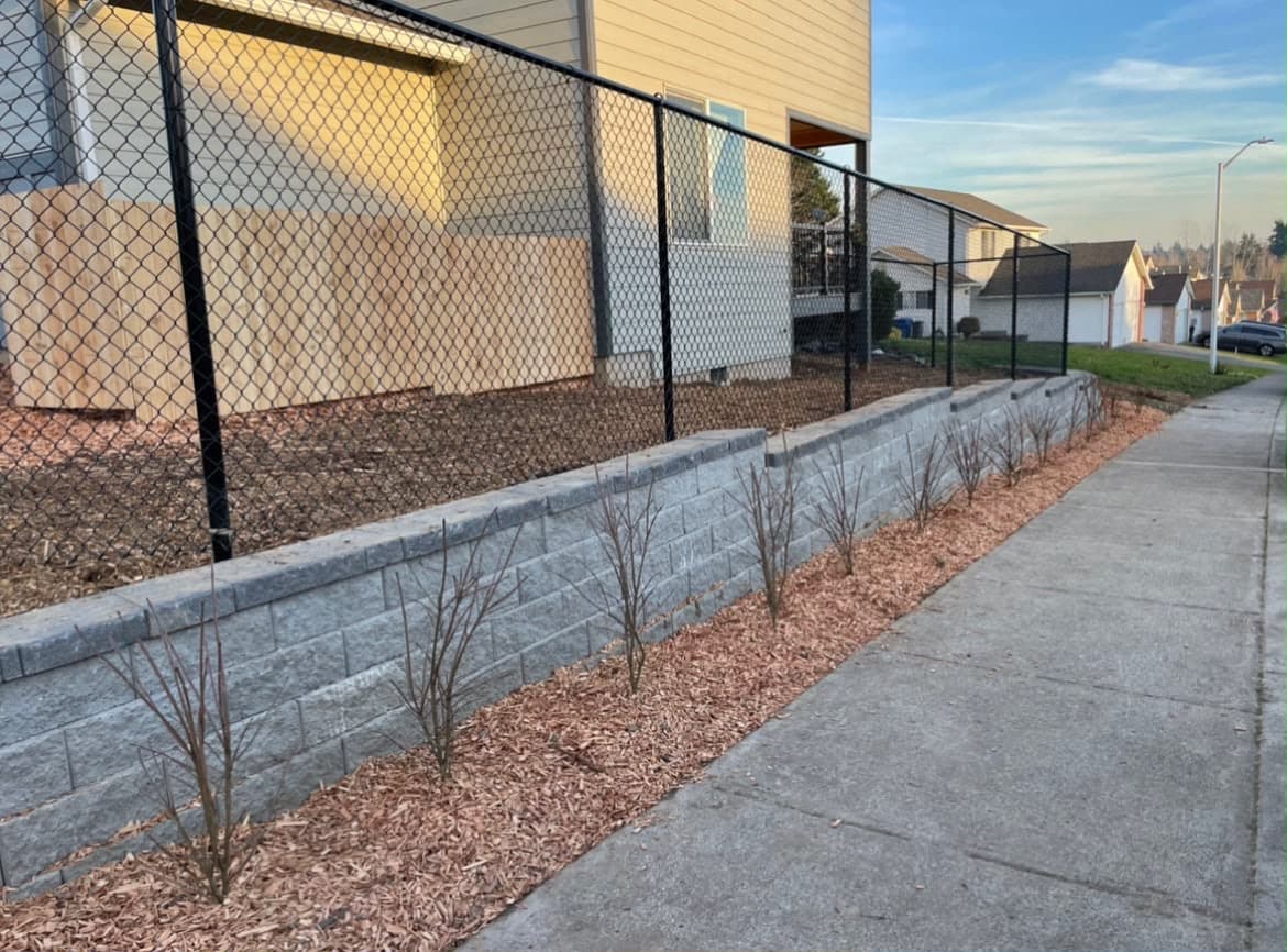 Grey segmental block retaining wall running along a public sidewalk with a fresh mulch bed and new shrub plantings, built by GI Landscape