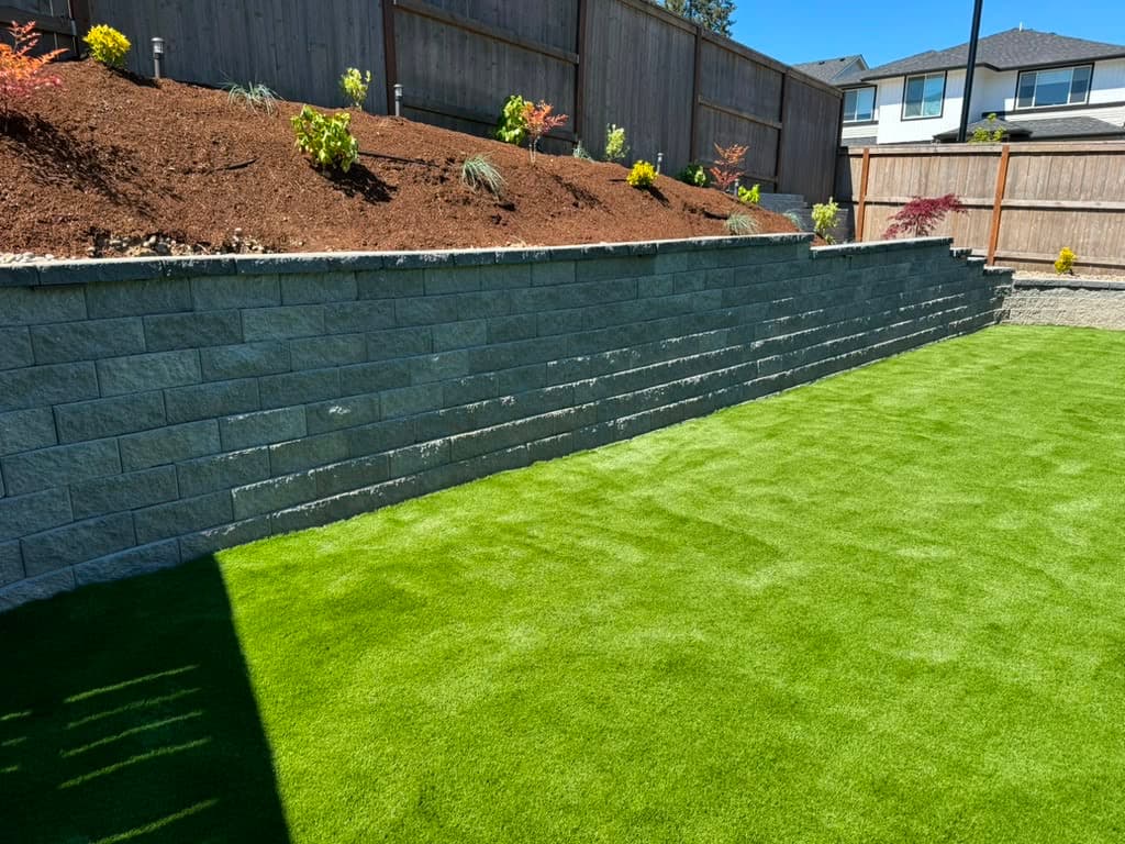 Charcoal segmental retaining wall with capstones holding back a planted slope above an artificial turf lawn, by GI Landscape