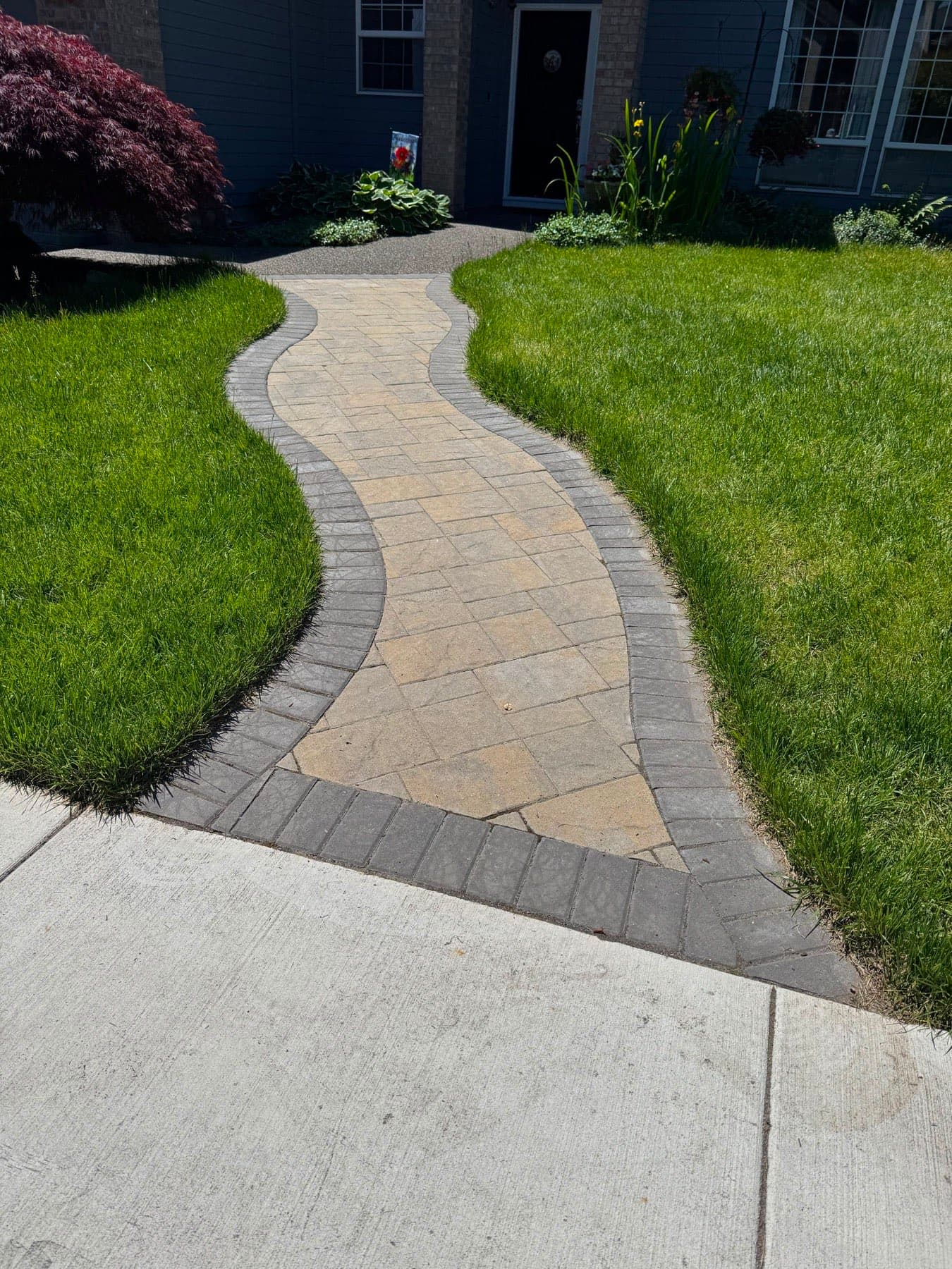 Curving tan paver front walkway with a charcoal soldier-course border, set through a green lawn to the front door, by GI Landscape