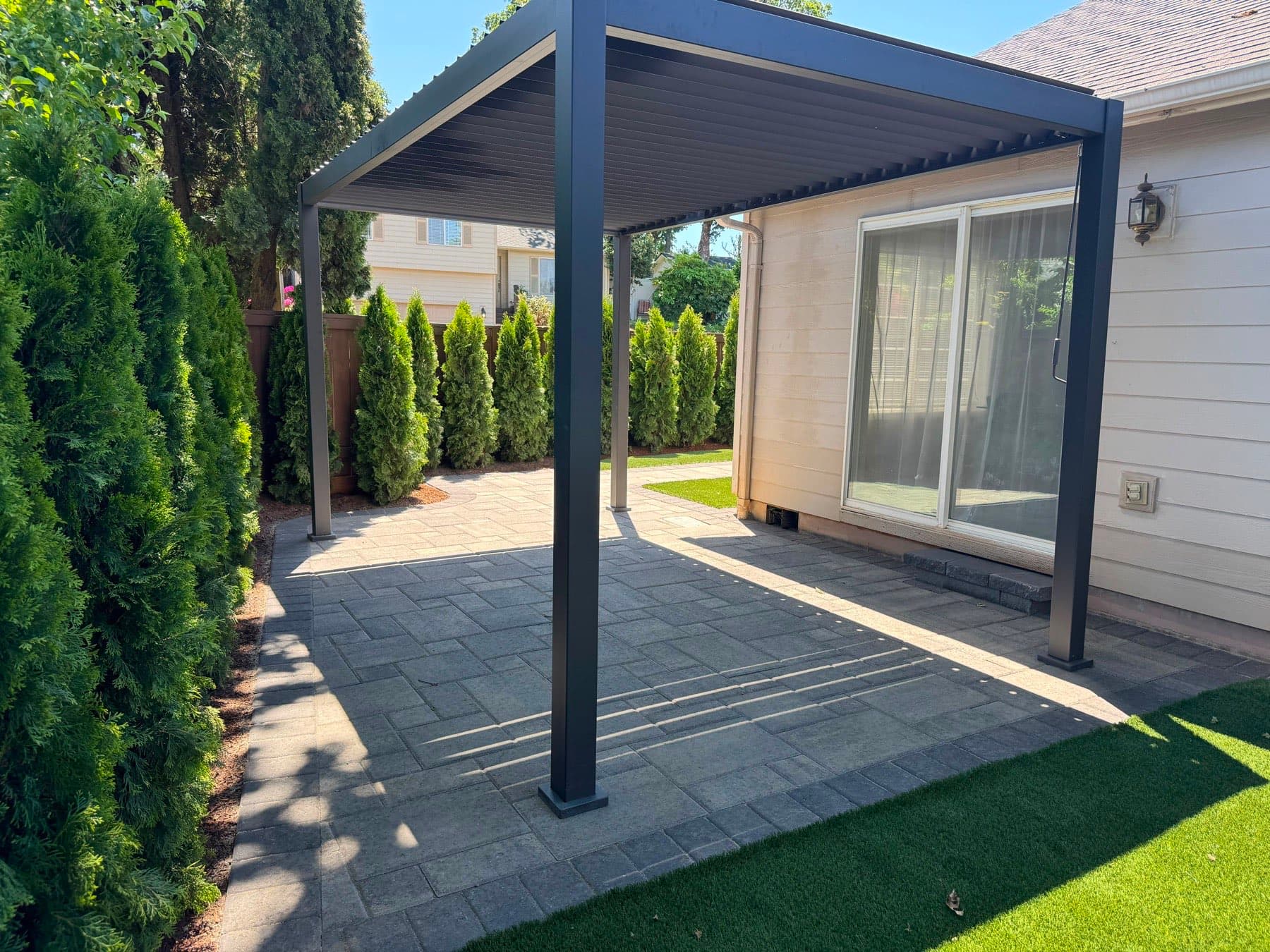 Modern black louvered pergola over a grey paver patio beside an artificial turf lawn and arborvitae screen, by GI Landscape