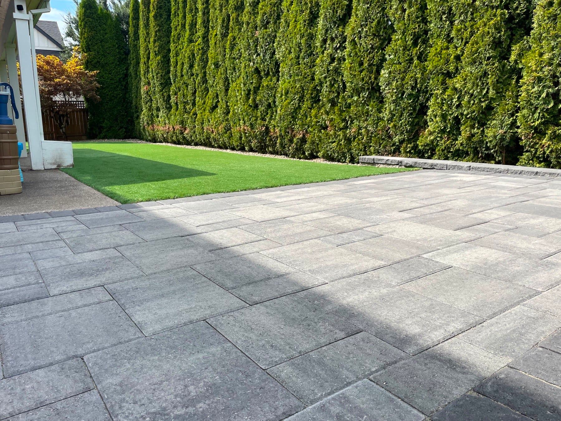 Wide grey paver patio meeting an artificial turf strip along a mature arborvitae hedge with a low block wall, by GI Landscape