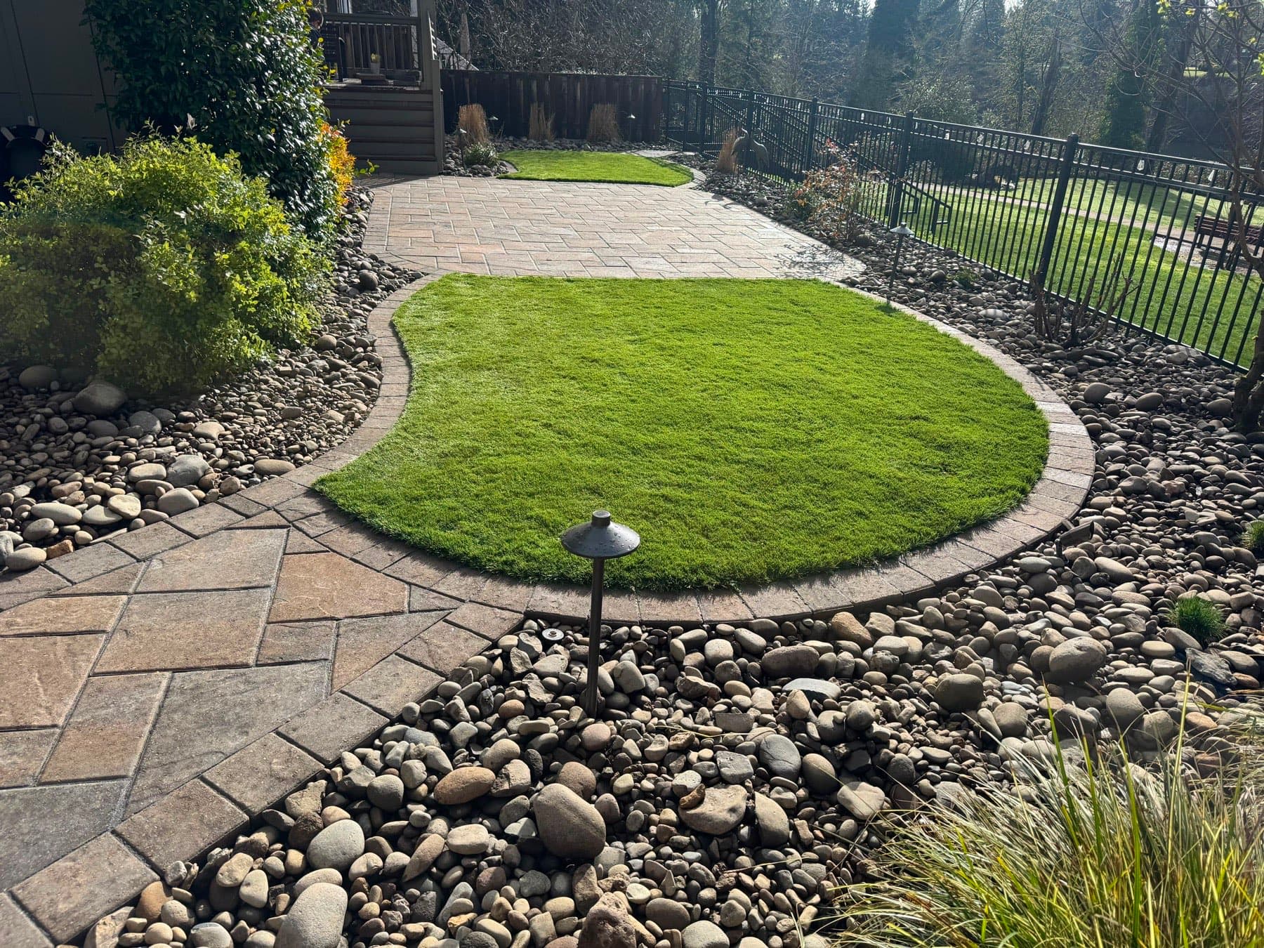 Curved paver patio wrapping an oval artificial turf lawn edged in paver banding, with river rock beds and path lighting, by GI Landscape