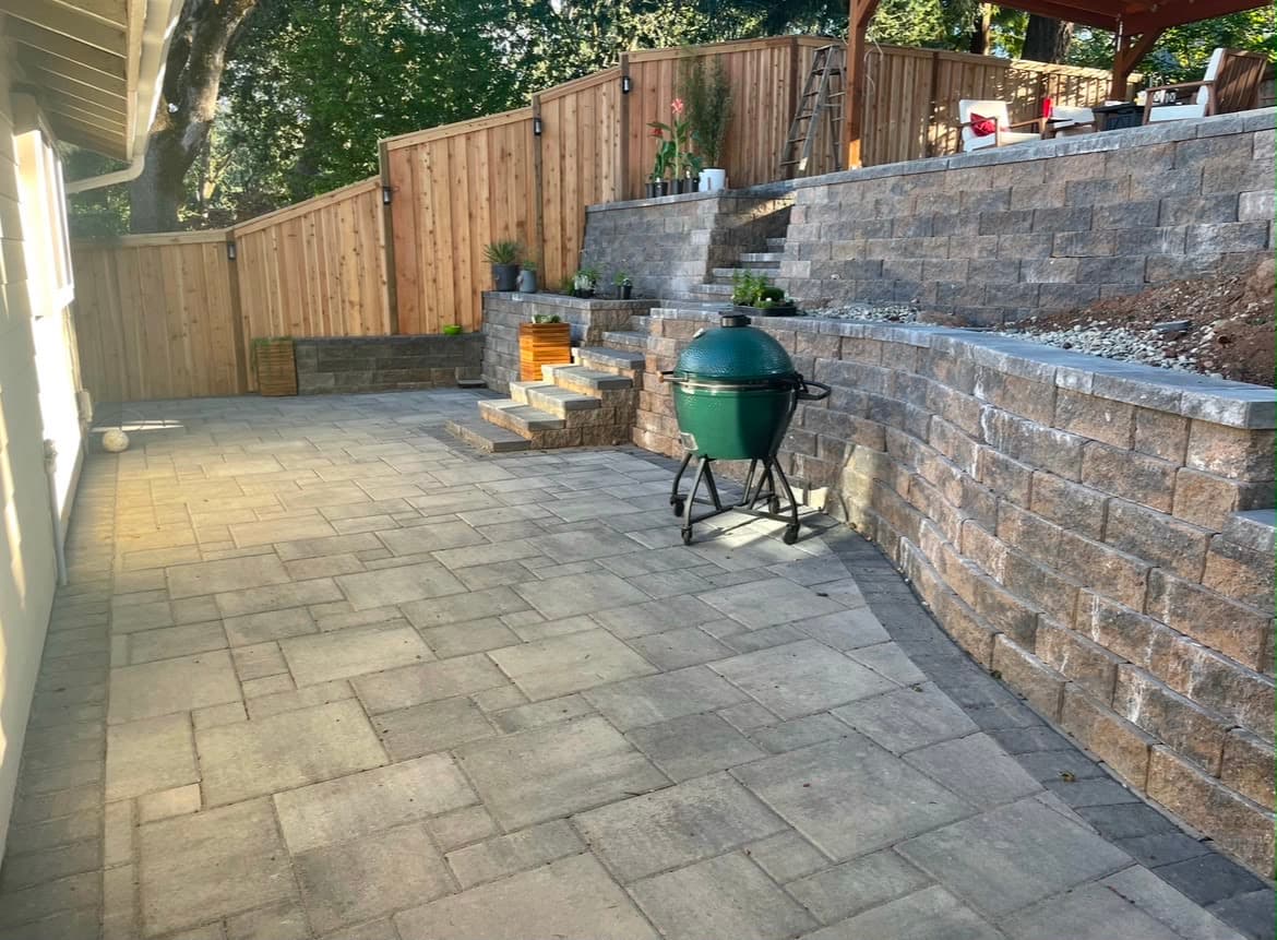 Paver patio below a tiered block retaining wall with integrated steps and a cedar fence above, by GI Landscape