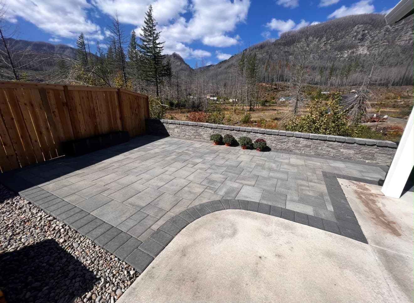 Grey paver patio with a charcoal border and stacked-stone seat wall, cedar fence and forested valley beyond, by GI Landscape