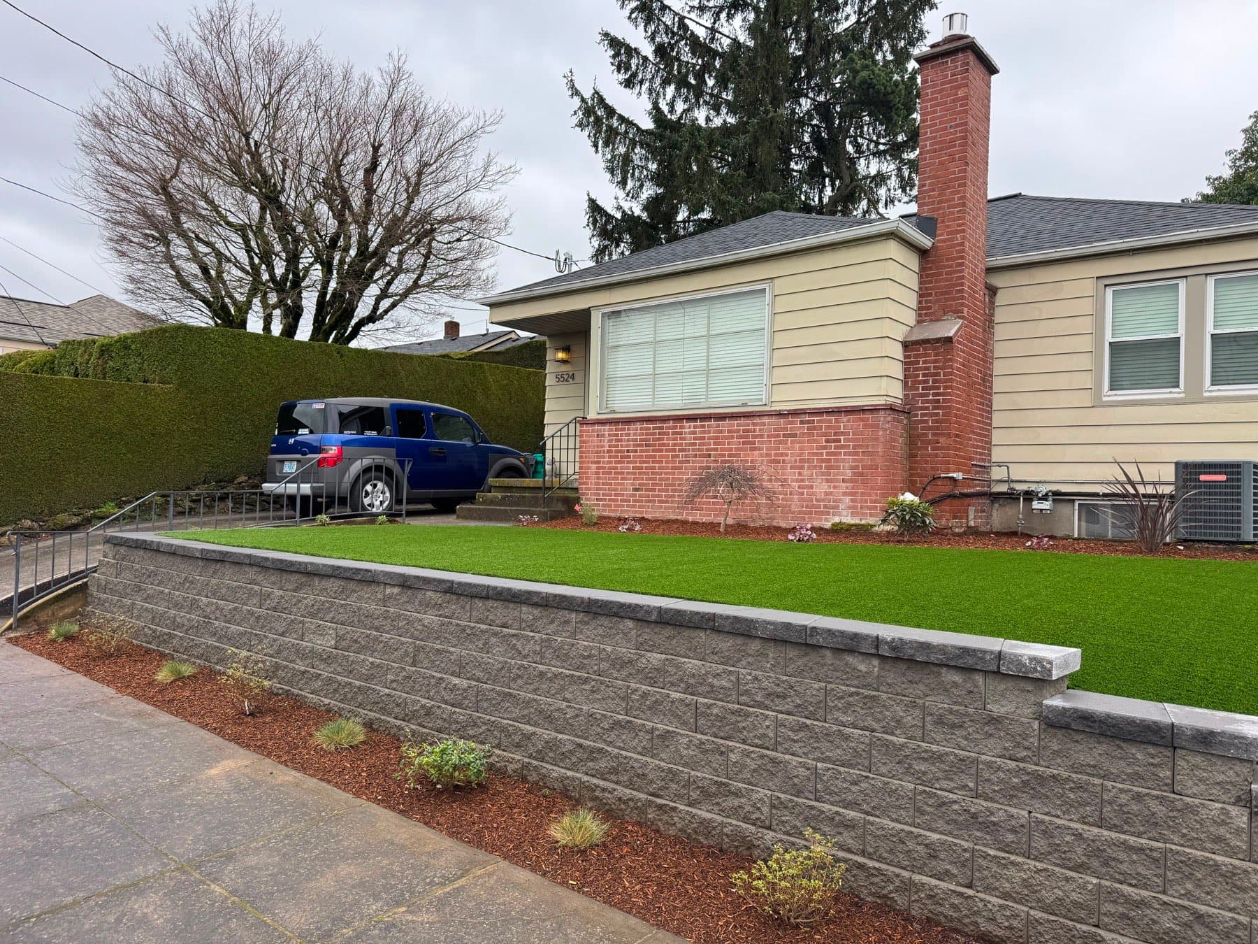 Grey block retaining wall with capstones lifting a front-yard artificial turf lawn above the driveway, by GI Landscape