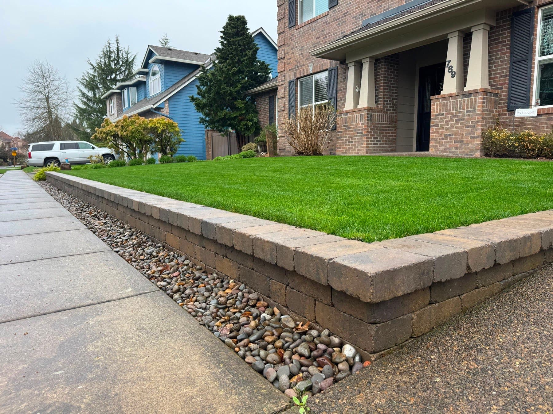 Tan block retaining wall with capstones and a river rock strip holding a level front lawn, by GI Landscape