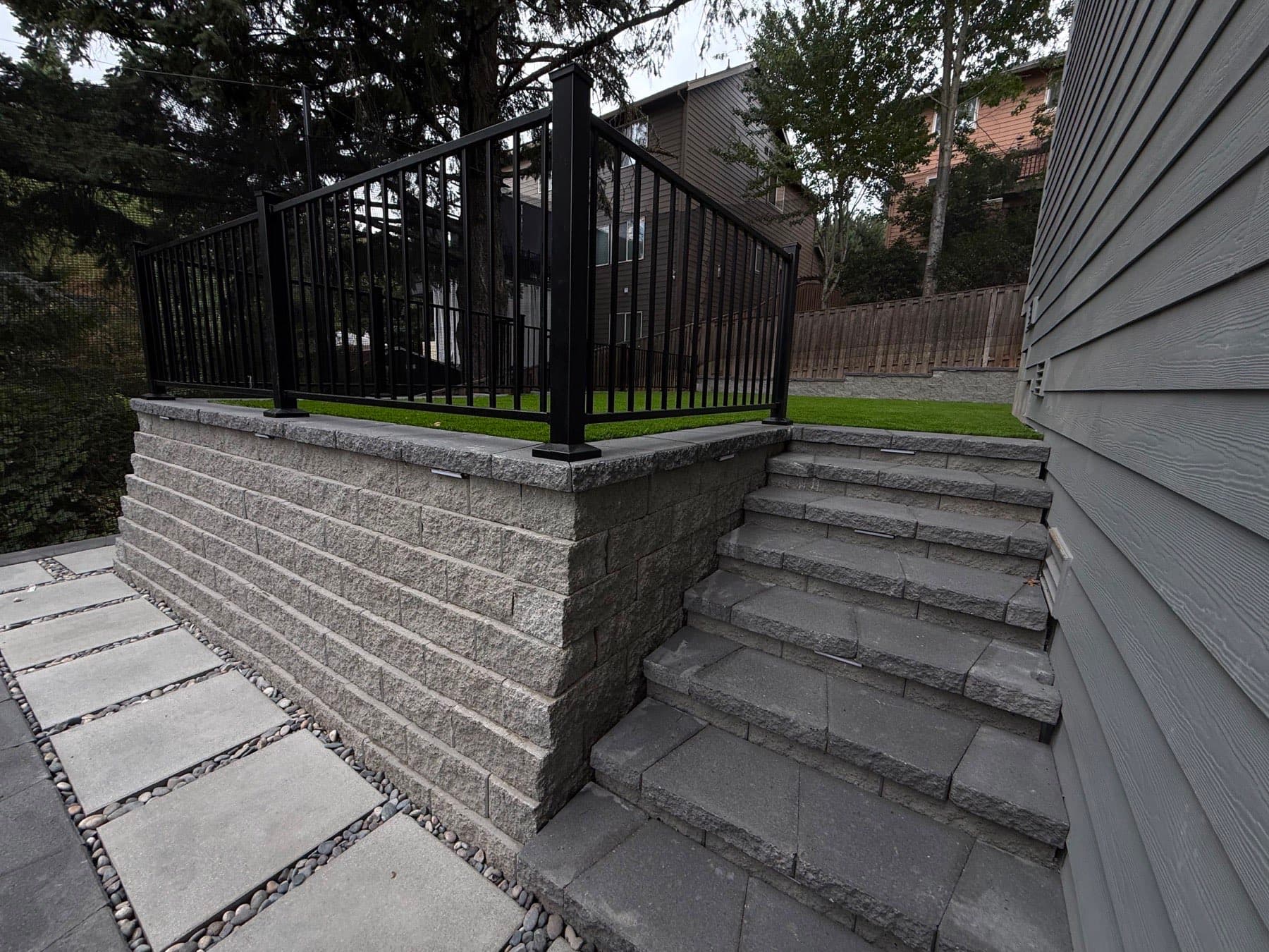 Block retaining wall with capstones and a black metal railing above lit paver steps and a river-rock jointed landing, by GI Landscape