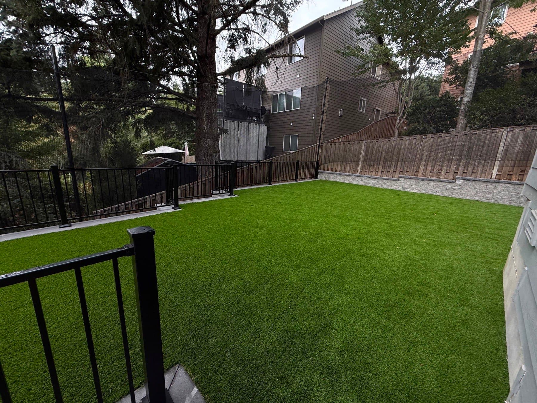 Raised artificial turf terrace enclosed by a black metal railing and block retaining wall, by GI Landscape