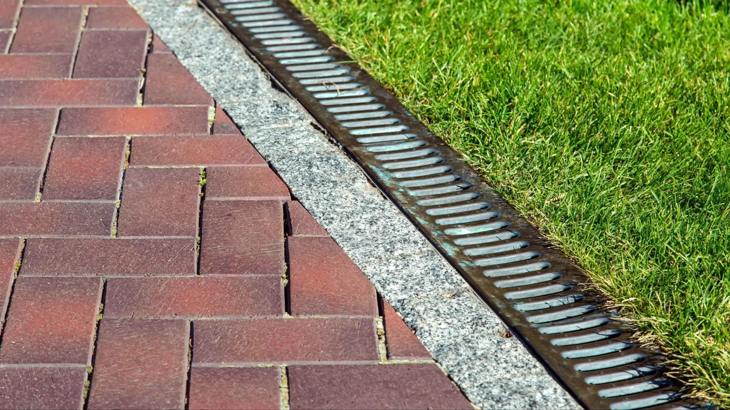 Channel drain with a metal grate set between a brick paver path and lawn, by GI Landscape