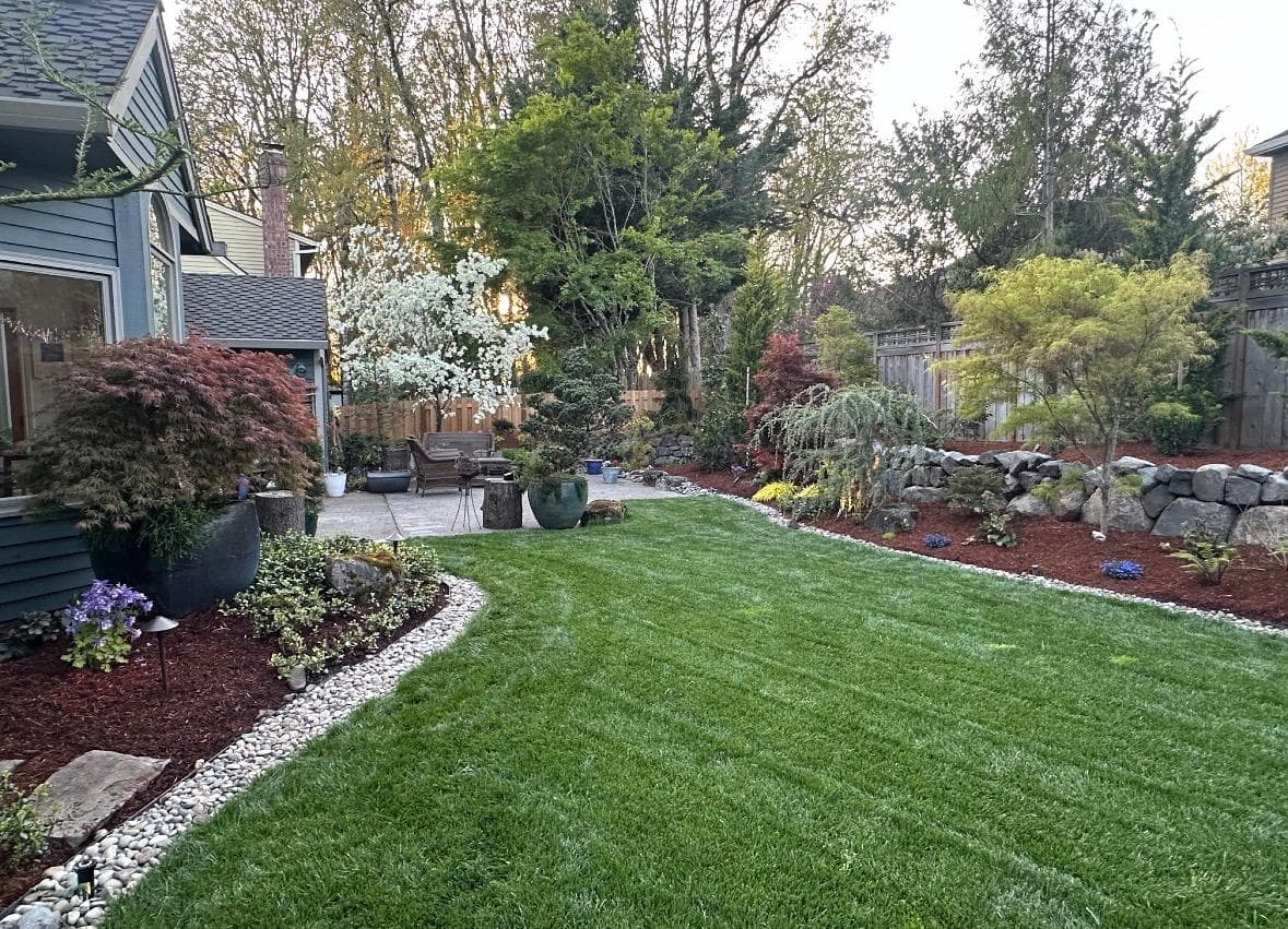 Backyard artificial-turf lawn with a white-pebble border and planting beds, by GI Landscape